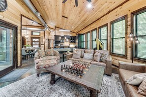 A cozy cabin interior with a wooden ceiling, a dining area, a living room with a chessboard, and large windows. - Hike, Soak & Play! Luxe Broken Bow Escape (Broken Bow)