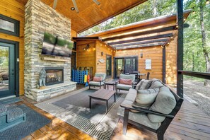 A covered outdoor seating area with a fireplace, wooden furniture, and a stone wall. - Hike, Soak & Play! Luxe Broken Bow Escape (Broken Bow)