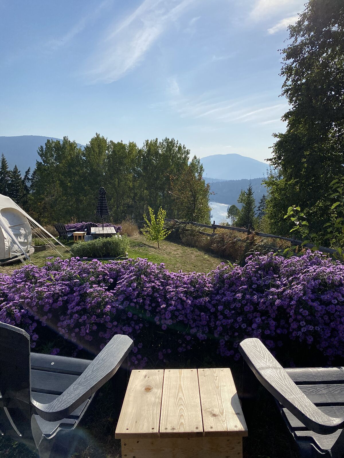 Una mesa de madera y dos sillas Adirondack en un jardín con flores moradas y vistas a un lago y montañas.