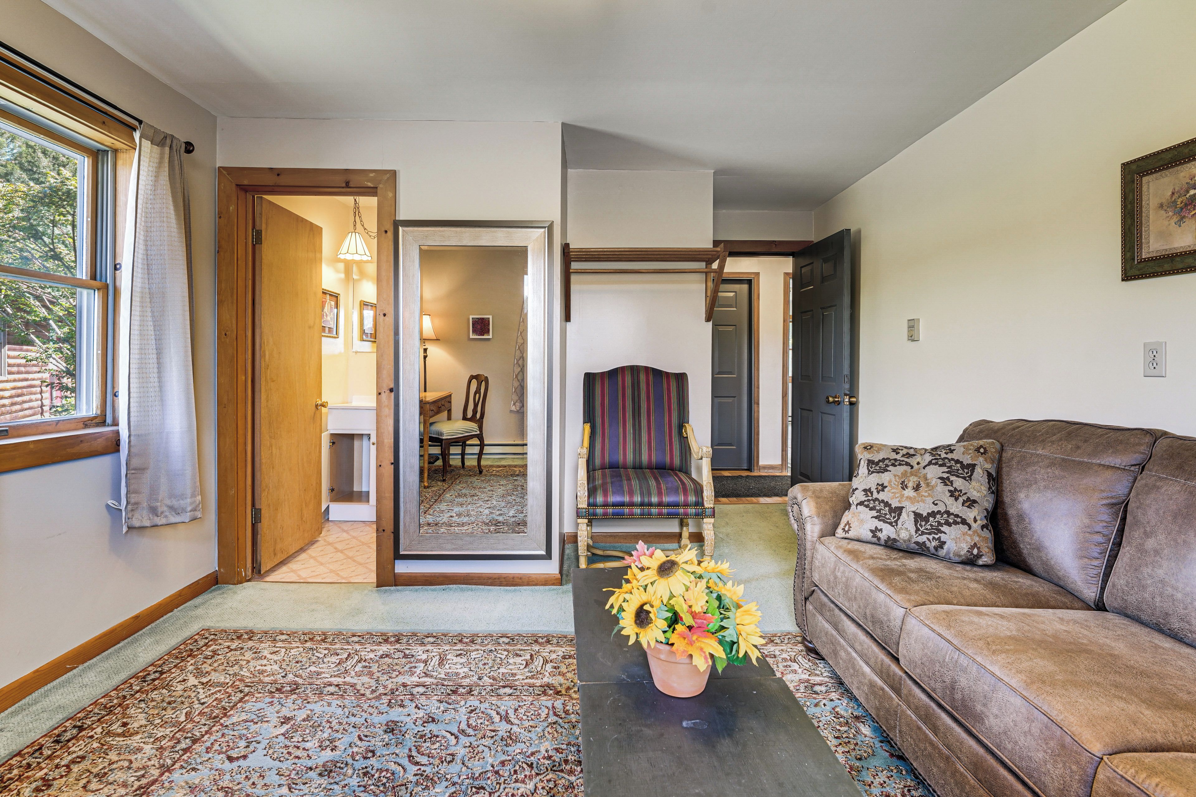 Una sala de estar con un sofá, una mesa de centro con un arreglo floral, un tapete estampado y vista a un comedor por una puerta abierta.