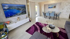 Ein modernes Wohnzimmer mit großem Flachbildfernseher, einer Couch, einer Essecke mit Stühlen und einem violetten Teppich.