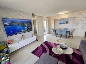 A modern living room with a large flat-screen TV, a sofa, a dining area with chairs, and a purple rug. - Lux Adriatica Apartments (Okrug)