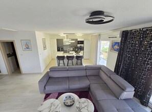 A modern living room with a grey sectional sofa, a dining area with bar stools, and a kitchen in the background. - Lux Adriatica Apartments (Okrug)