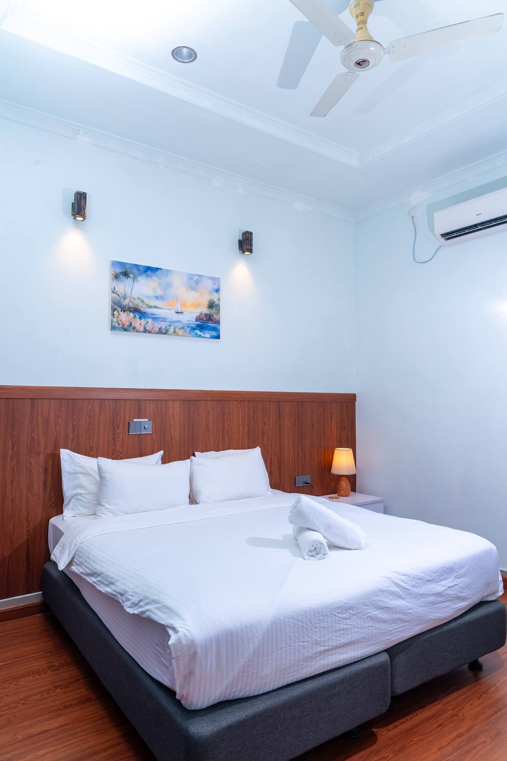 Habitación de hotel con cama, ventilador de techo, unidad de aire acondicionado y un cuadro en la pared.