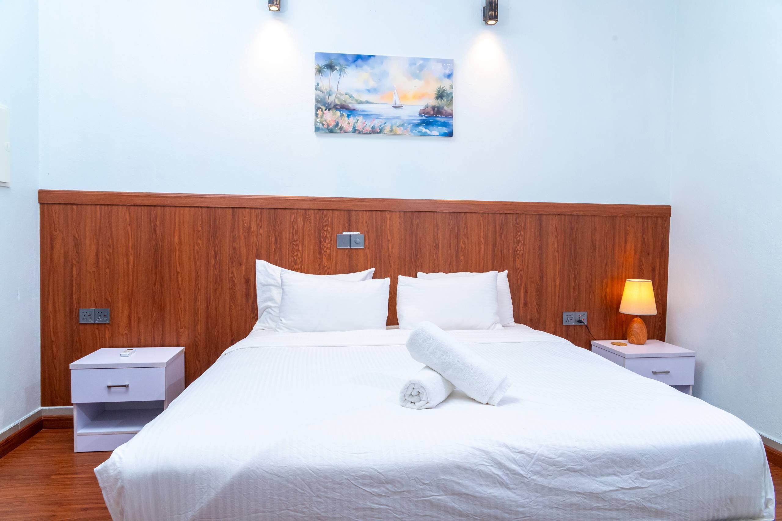 A hotel room with a wooden headboard, white bedding, a painting on the wall, and a bedside lamp.
