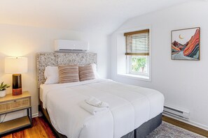 Un dormitorio con cama, mesita de noche con lámpara, aire acondicionado instalado en la pared, ventana con persianas y un cuadro en la pared.