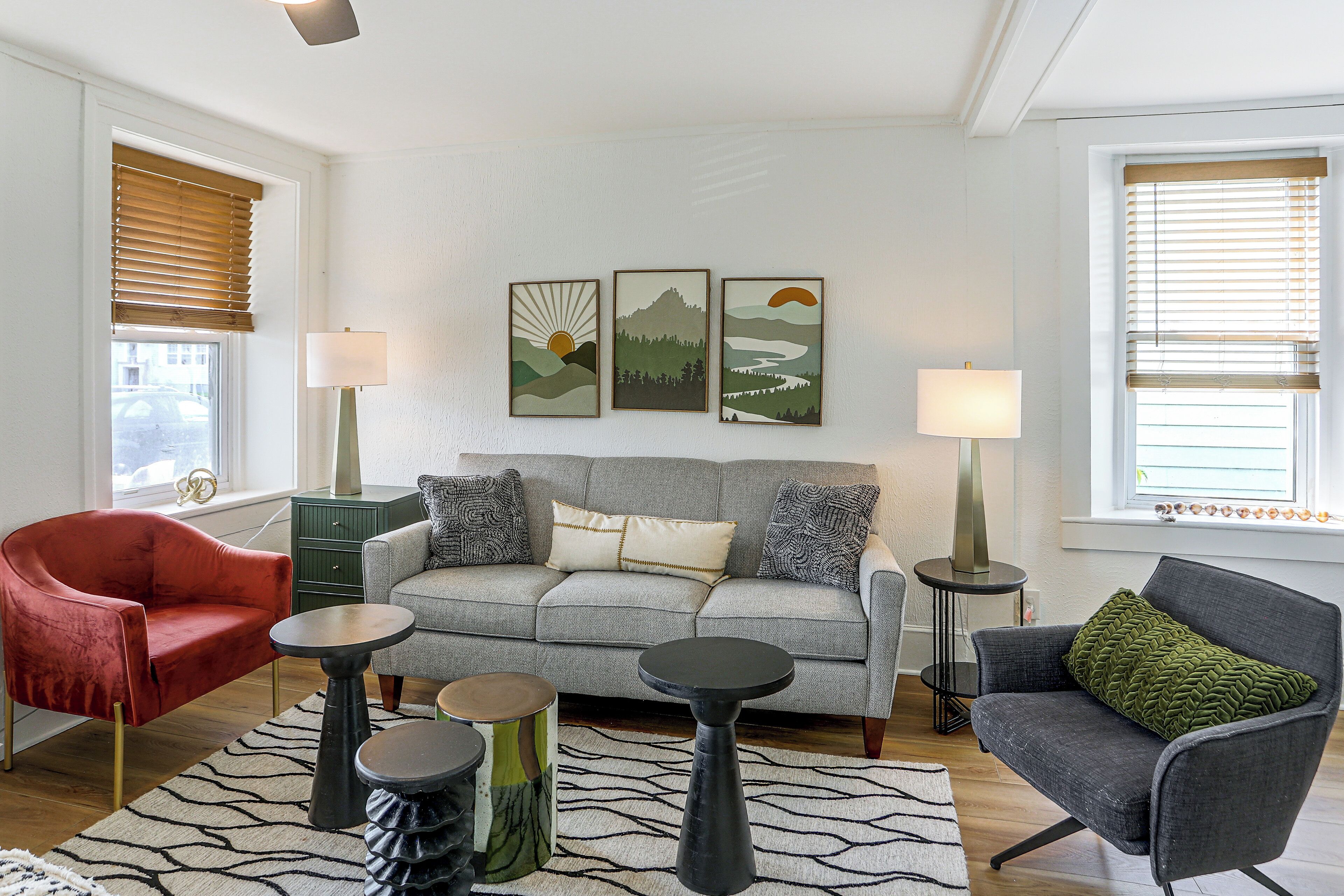 Un salon avec un canapé gris, un fauteuil rouge et un fauteuil vert. Trois œuvres d’art encadrées ornent le mur, une lampe se trouve sur une table d’appoint et un tapis recouvre le sol.