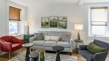 Un salon avec un canapé gris, un fauteuil rouge et un fauteuil vert. Trois œuvres d’art encadrées ornent le mur, une lampe se trouve sur une table d’appoint et un tapis recouvre le sol.