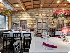 Ein Restaurantinneres mit Backsteinwänden, Holzbalken und einer rustikalen Tür. Die Tische sind mit weißen Tischdecken und roten Servietten gedeckt.