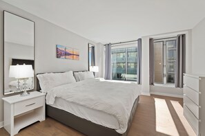 A modern bedroom with a large bed, a nightstand, a mirror, and a window with curtains. - SW DC Waterfront Apt at The Wharf | City Views (Washington)