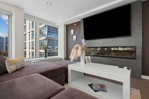 A modern living room with a large flat-screen TV mounted on the wall, a sofa, a coffee table, and a fireplace. - SW DC Waterfront Apt at The Wharf | City Views (Washington)