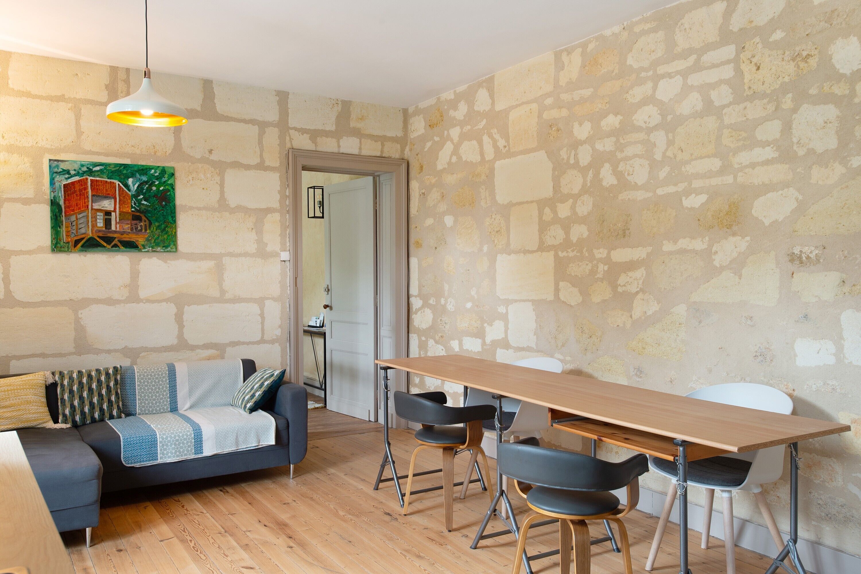 Une pièce comprenant une table à manger en bois, deux chaises, un canapé avec des coussins à motifs, un luminaire suspendu et un mur en pierre.