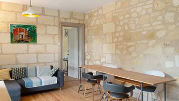 A room with a wooden dining table, two chairs, a sofa with patterned cushions, a hanging light fixture, and a stone wall.