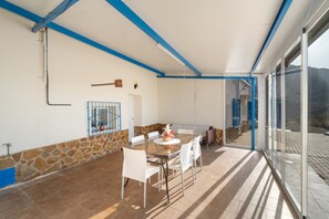 Un patio techado con una mesa y sillas, pared de piedra y vistas a través de puertas de vidrio.