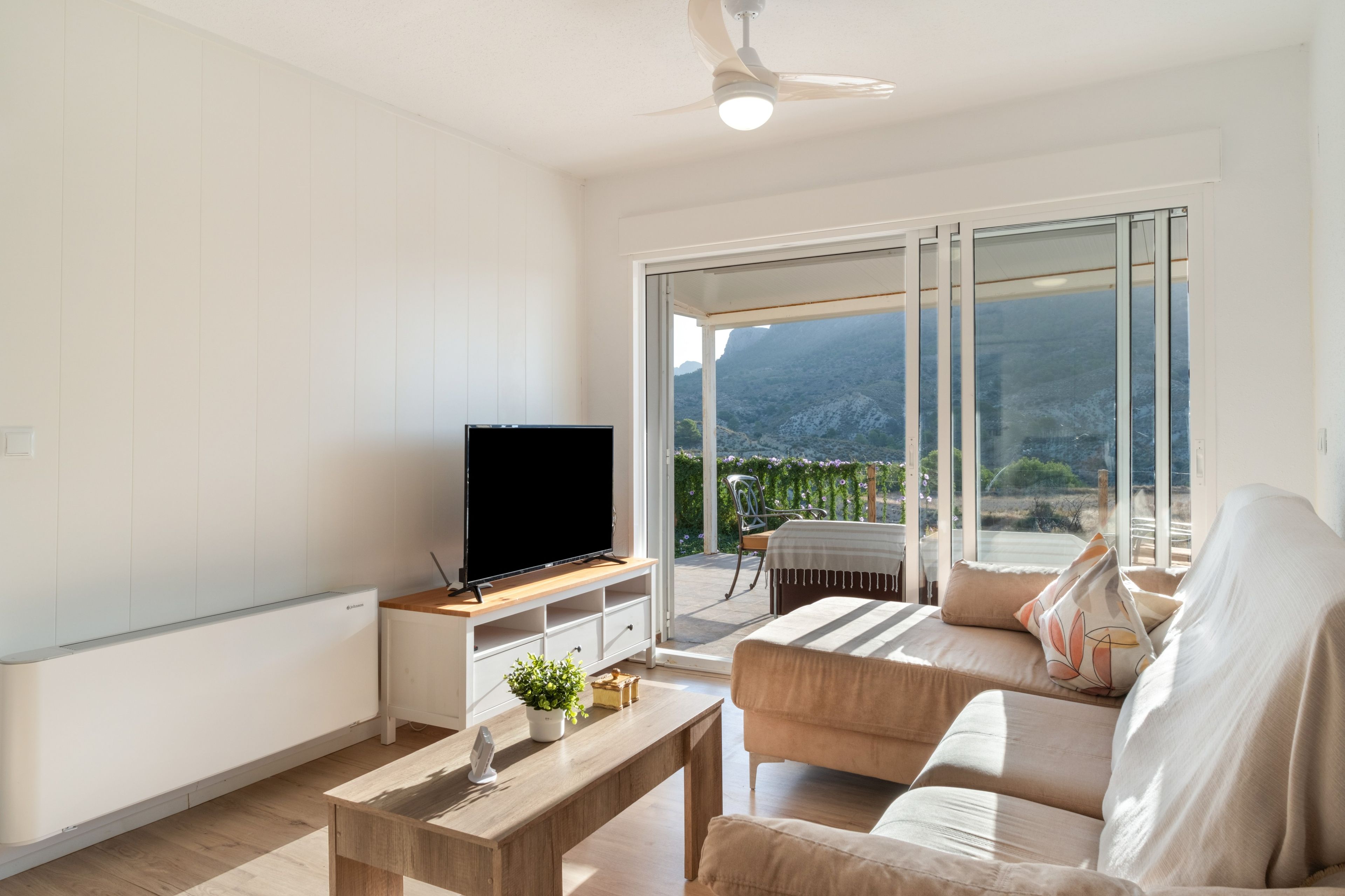 Ein modernes Wohnzimmer mit einem Flachbildfernseher auf einem Holzboden, einer Couch, einem Couchtisch und einem Balkon mit Bergblick.