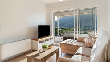 Ein modernes Wohnzimmer mit einem Flachbildfernseher auf einem Holzboden, einer Couch, einem Couchtisch und einem Balkon mit Bergblick.