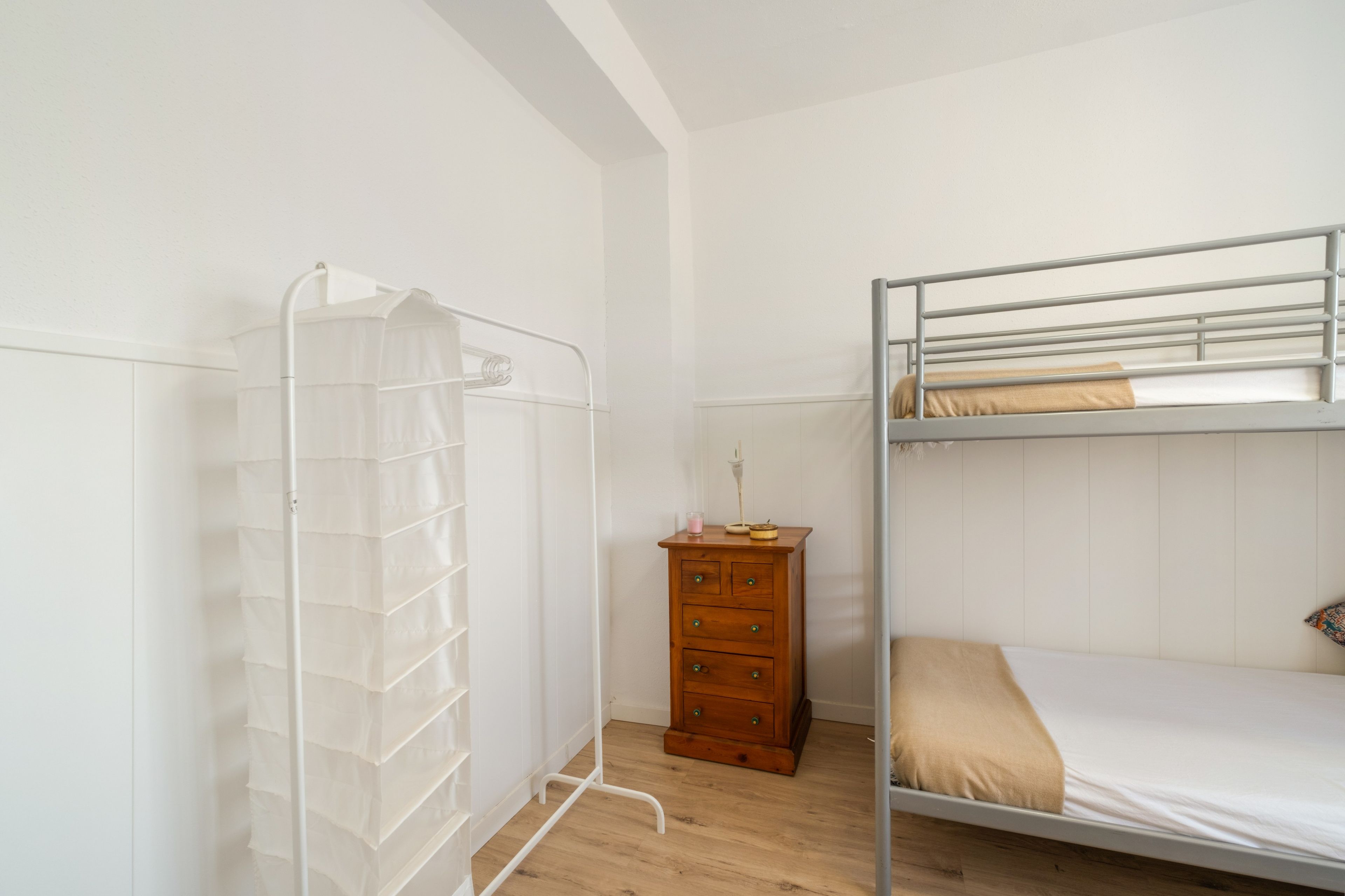 Een stapelbed met een houten dressoir en een witte rek voor het drogen van kleding in een kamer met witte muren en houten vloeren.