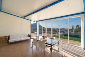 Una habitación iluminada por el sol con una mesa y sillas de vidrio, un sofá y vistas a una piscina y montañas.