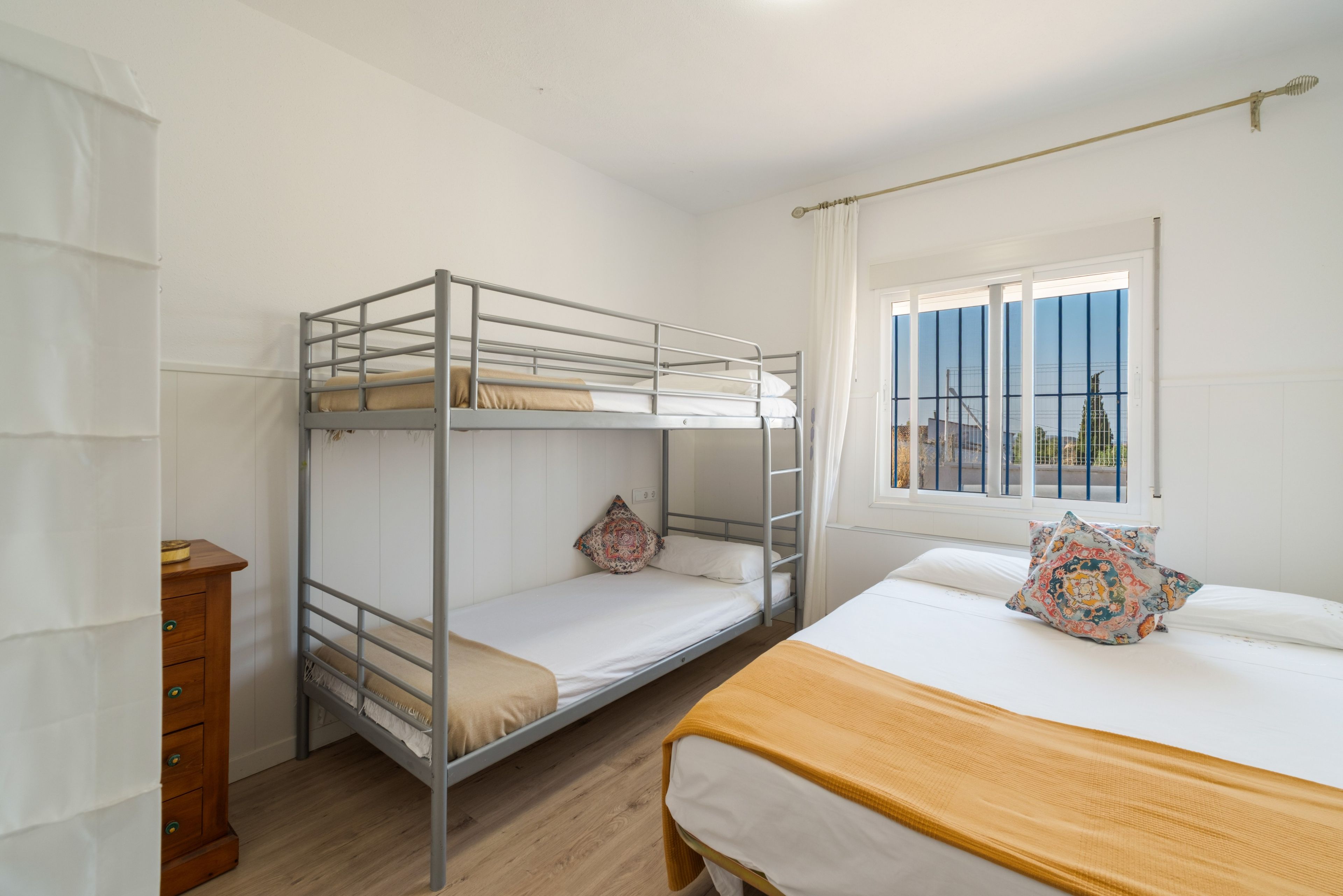 Habitación tipo dormitorio con literas, un aparador de madera, una ventana con cortinas y una cama con almohada estampada.