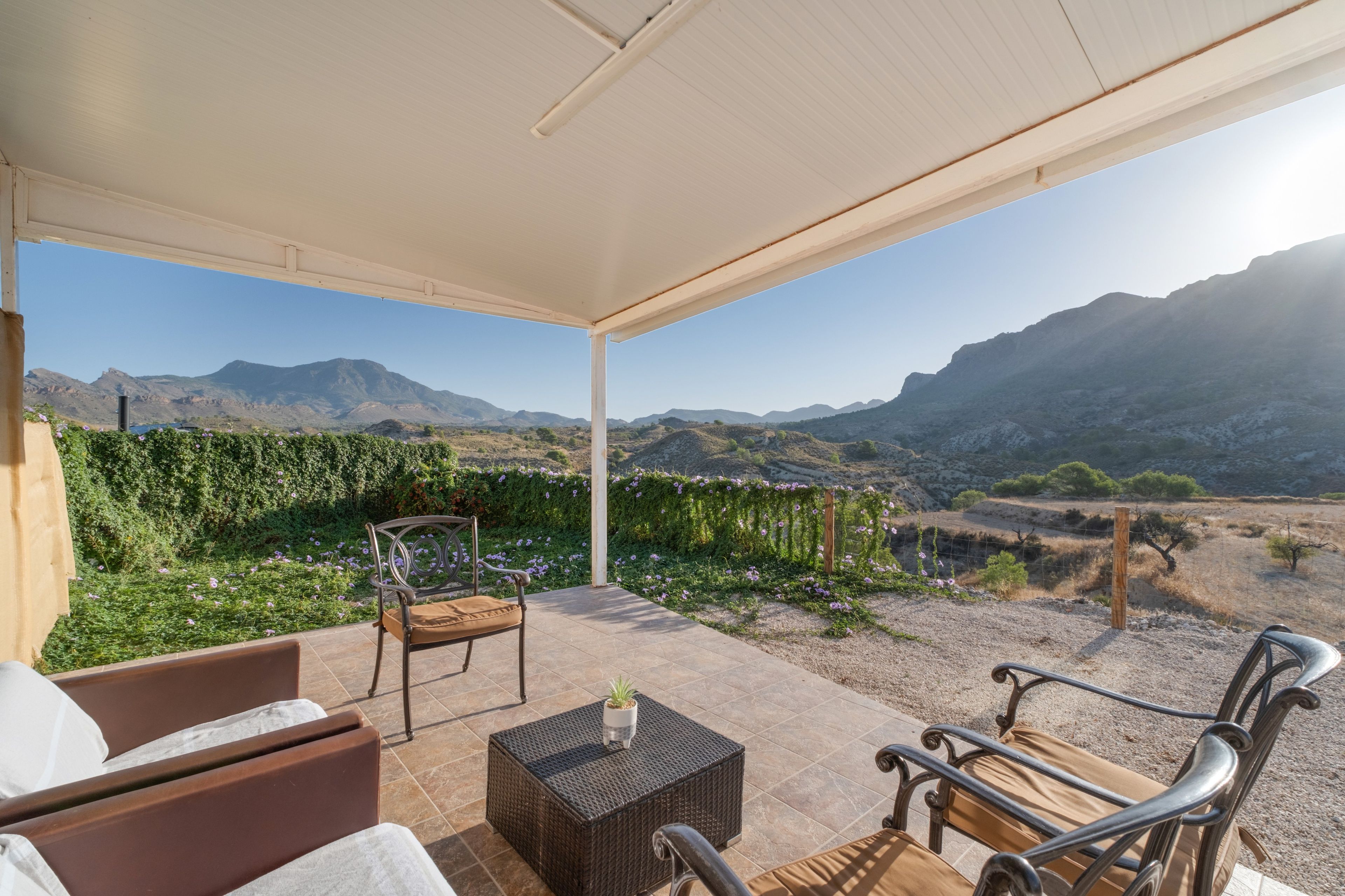 Eine überdachte Terrasse mit Gartenmöbeln, Blick auf Berge und einen Garten.