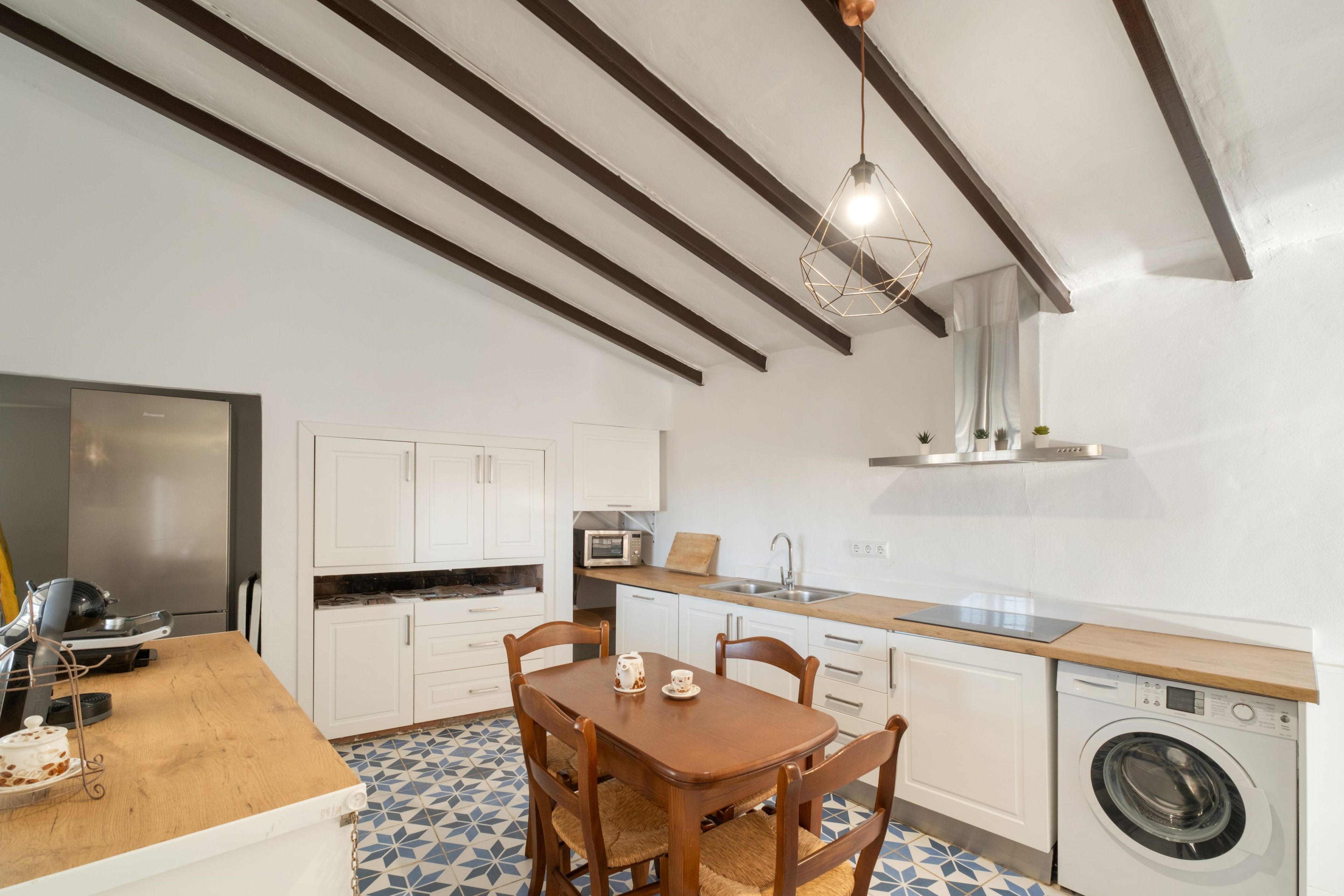 Une cuisine compacte avec une table à manger en bois, des placards blancs, un lave-linge encastré et un luminaire suspendu.