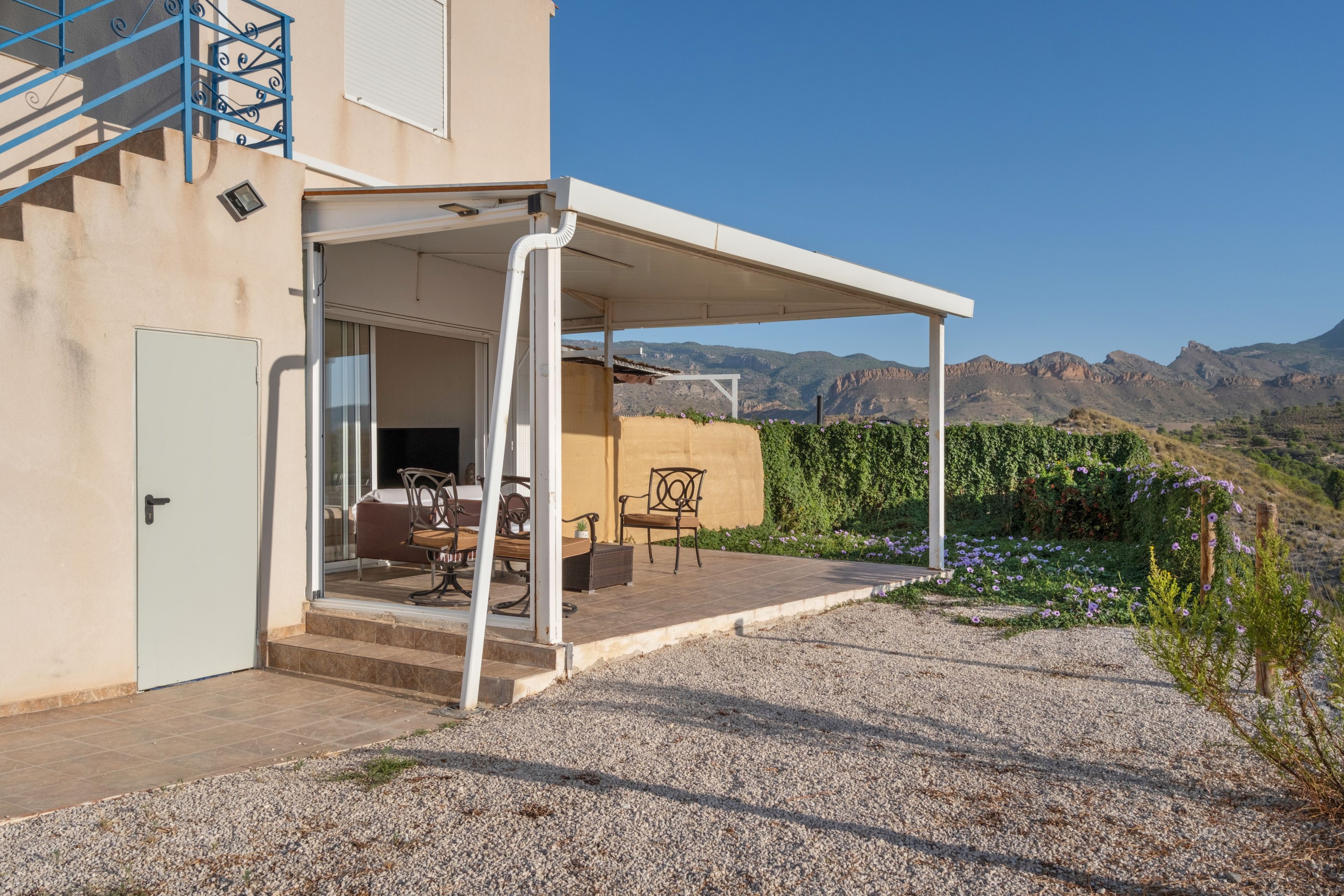 Un patio cubierto con mobiliario exterior, una puerta que da a un espacio interior y una vista de montañas al fondo.