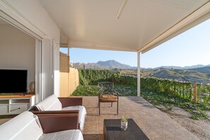 A covered outdoor seating area with a TV, a sofa, and a chair, overlooking a mountainous landscape.