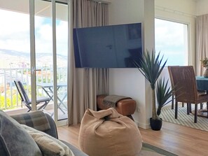 A living room with a large TV, a sofa, a coffee table, and a view of a cityscape through a balcony. - Seascape Penthouse - Wake Up to the Atlantic | Oceanfront Penthouse (Funchal)