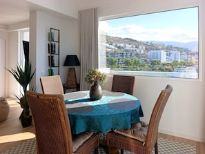 A dining area with a table, chairs, a vase with flowers, a bookshelf, and a view of a cityscape through a window. - Seascape Penthouse - Wake Up to the Atlantic | Oceanfront Penthouse (Funchal)