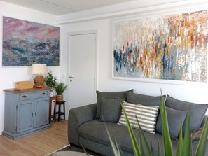 Een woonkamer met een grijze bank, een houten kast, een tafeltje met een lamp en twee potplanten.
