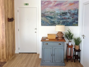 A room with a wooden floor, a white door, a grey cabinet with drawers, a painting on the wall, and a lamp on the cabinet. - Seascape Penthouse - Wake Up to the Atlantic | Oceanfront Penthouse (Funchal)