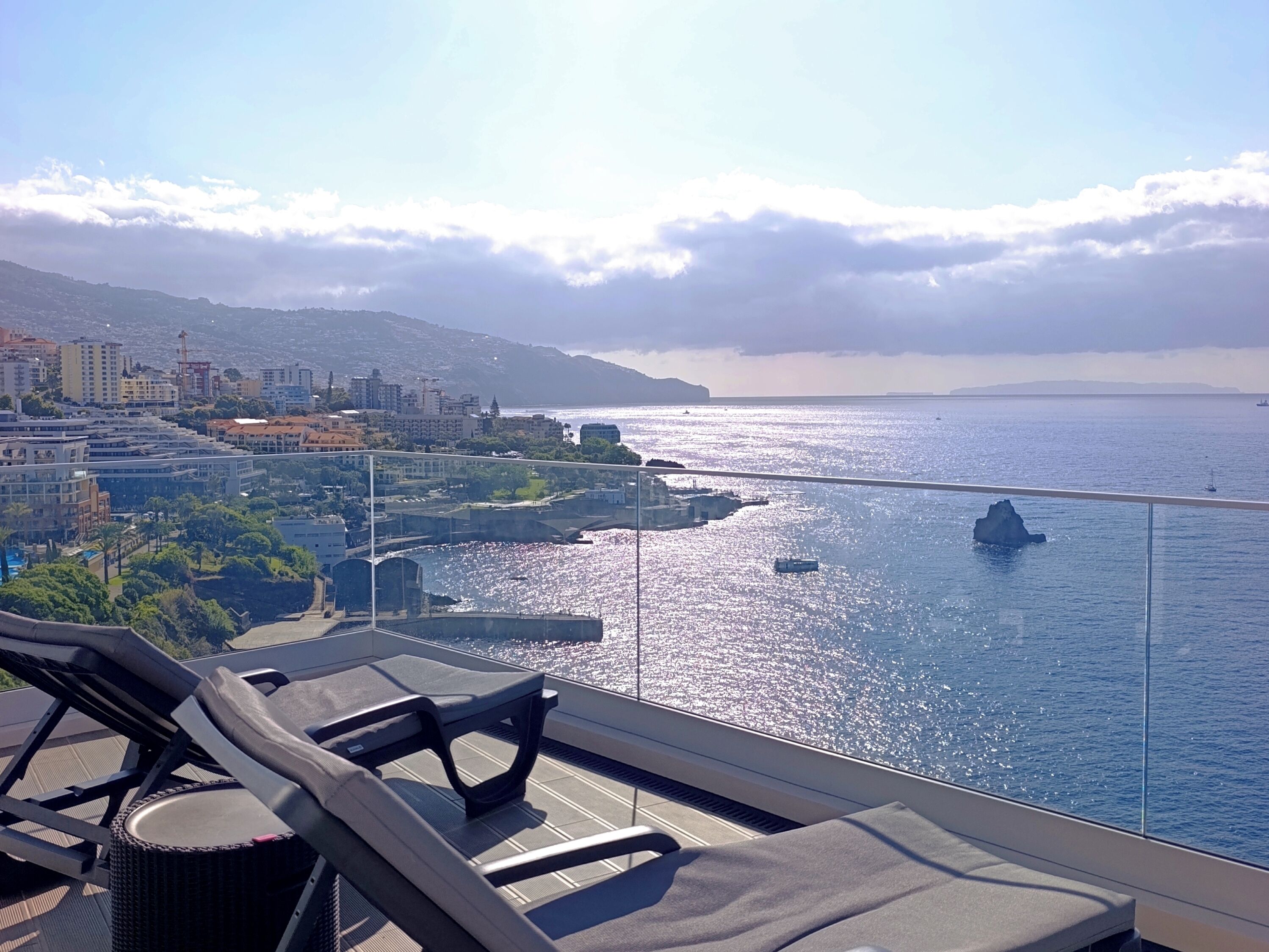 A balcony with lounge chairs overlooking a coastal cityscape.