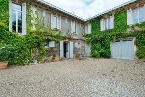 En gård med grusbelægning, et lille bord med stole og en dør, der fører til en bygning med slyngplanter på væggene og vinduer.