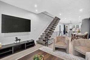 A modern living room with a flat-screen TV, a wooden entertainment unit, a staircase, and a dining area with a table and chairs.