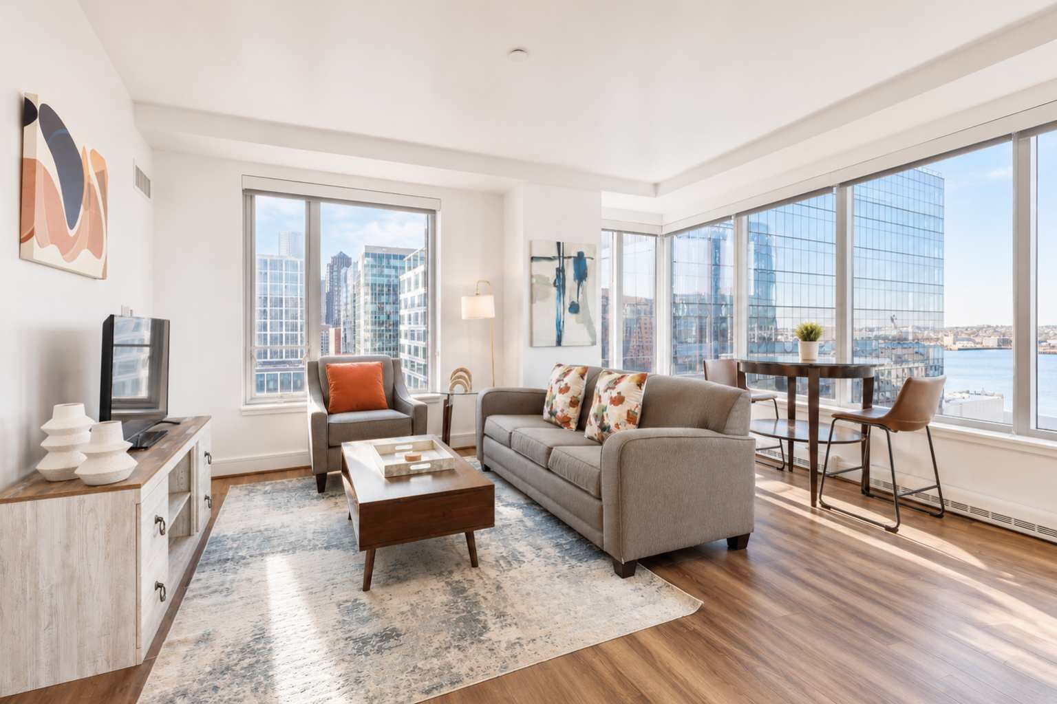Una sala de estar moderna con sofá, mesa de centro y televisor sobre un mueble de madera. Amplias ventanas ofrecen vistas a la ciudad.