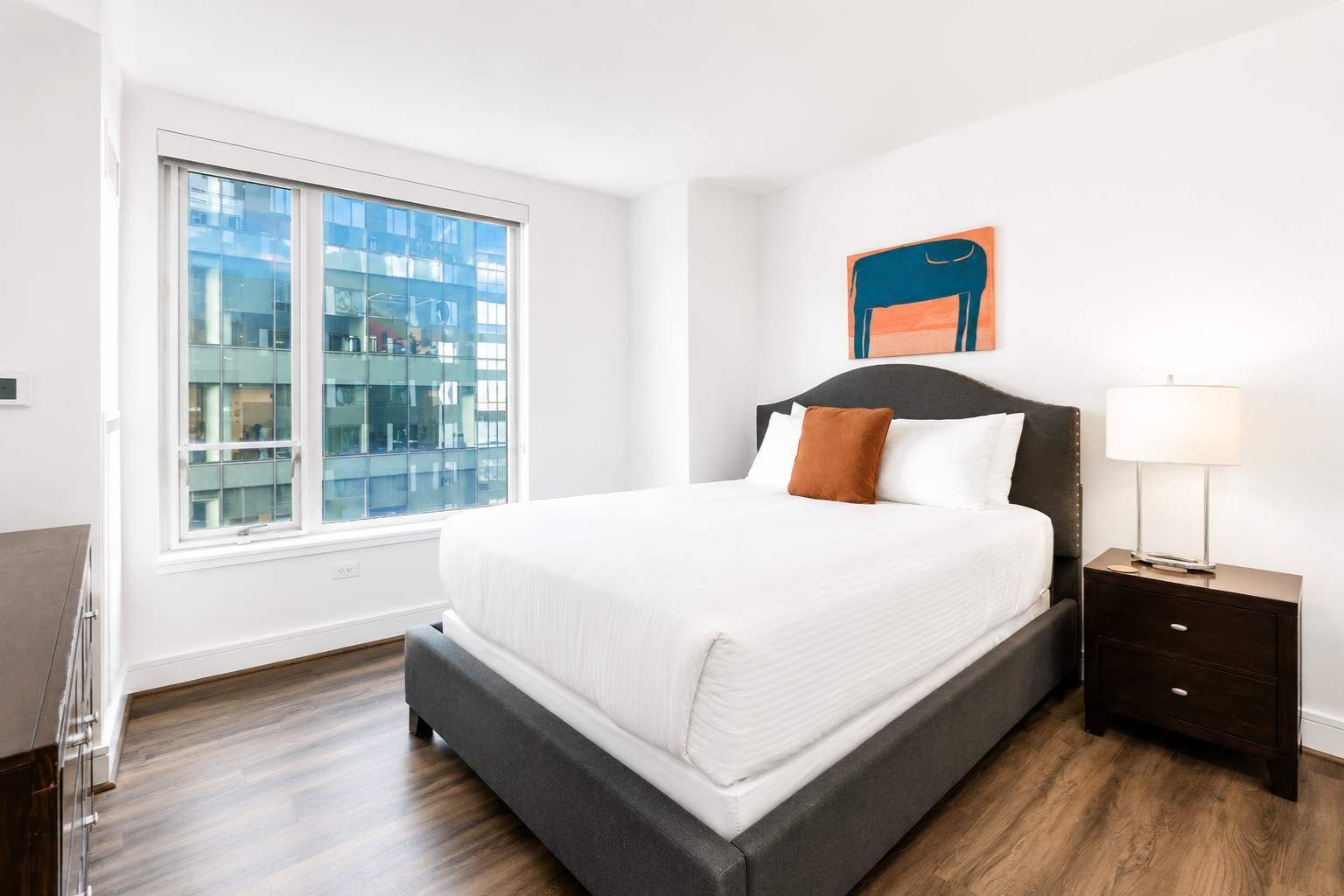 Een moderne hotelkamer met een groot raam, een bed met witte beddengoed, een nachtkastje met een lamp en een aan de muur hangend kunstwerk.