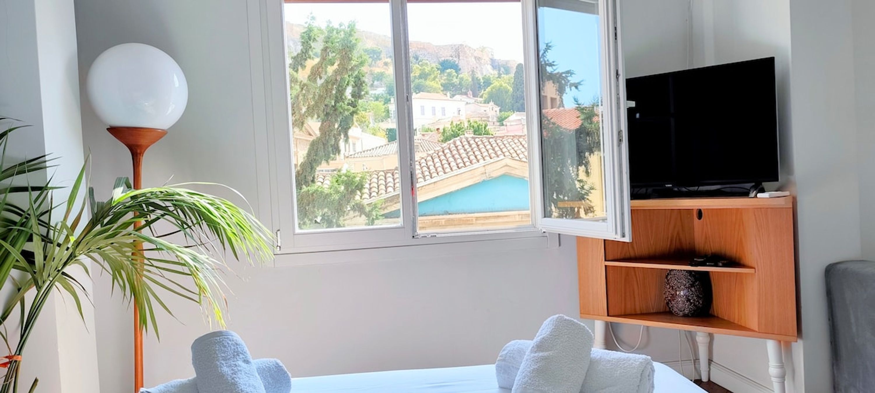 Ein Zimmer mit einem Bett, einem Fernseher auf einem Holzboden, einem großen Fenster mit Blick auf Gebäude und einer Zimmerpflanze.