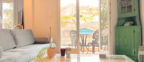 A living room with a sofa, a table with a plant and a cup, a green cabinet, and a view of a balcony with outdoor furniture.