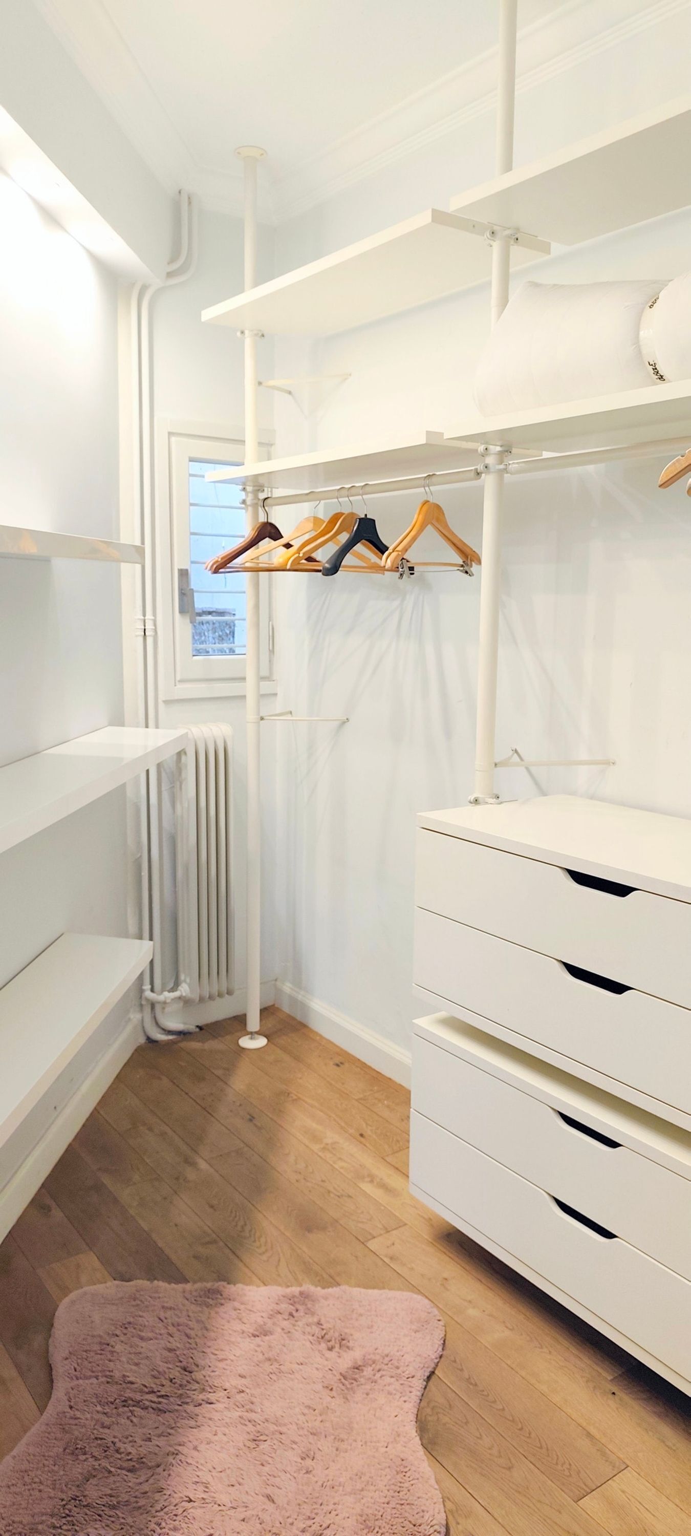 Een inloopkast met witte planken, een radiator en een roze vloerkleed.
