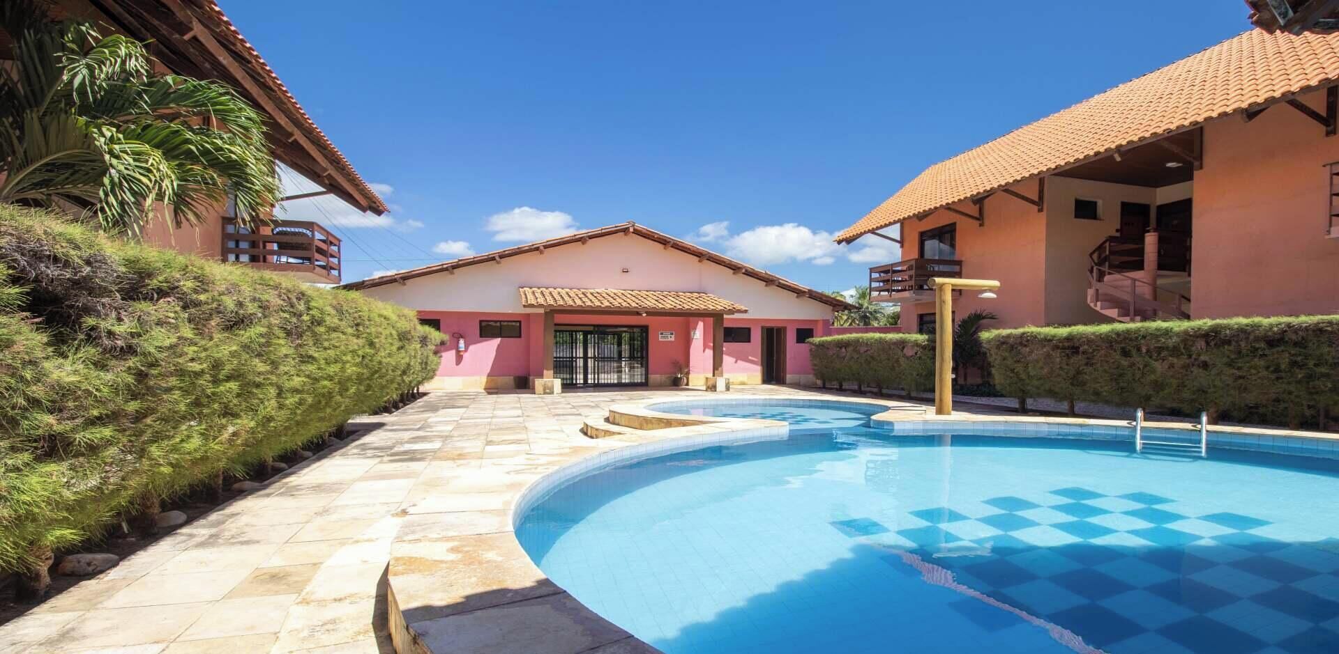Uma piscina com padrão quadriculado, um edifício com fachada rosa e um céu azul límpido.