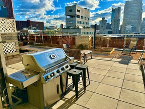 An outdoor patio with a barbecue grill, seating area, and a cityscape view in the background. - Luxury in Little Italy 2 Bedroom - Private Balcony (New York)