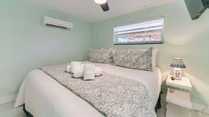 A bedroom with a bed, a ceiling fan, an air conditioning unit, a window with a view, and a bedside table with a lamp. - Sunshine Nook - 1/2 Block to the Beach (Fort Myers Beach)