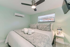 A bedroom with a bed, a ceiling fan, an air conditioning unit, a window with a view, and a bedside table with a lamp. - Sunshine Nook - 1/2 Block to the Beach (Fort Myers Beach)