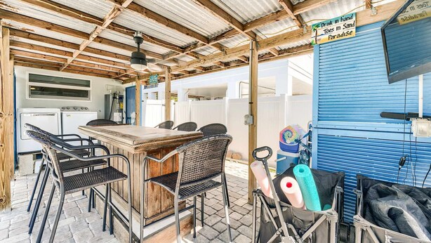 Cocina rústica al aire libre con barra, asientos y un letrero que indica bebidas en mano, paraíso de arena.