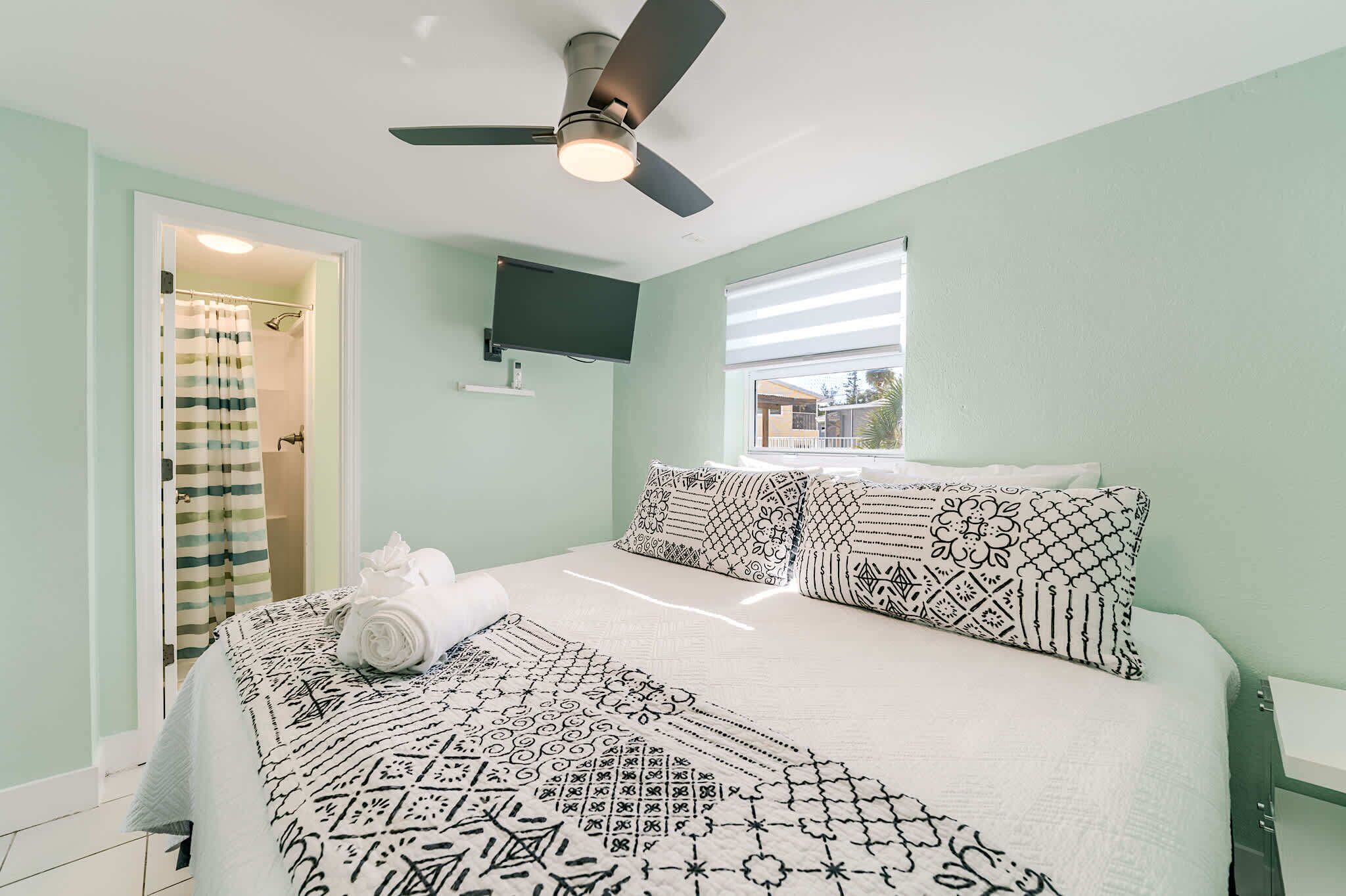 A bedroom with a bed, a ceiling fan, a TV mounted on the wall, and a window with a view.