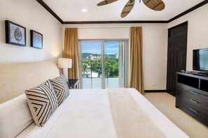 A hotel room with a bed, a television, a desk with a lamp, and a view of the outdoors. - Oceanfront 3BR | Sunset Views | Diamante del Sol (Jacó)
