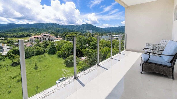 Uma varanda com vista para uma paisagem verde e montanhas, uma cadeira de vime e um corrimão.