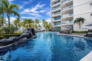 A swimming pool with a rock feature, surrounded by palm trees and a modern building with balconies. - Oceanfront 3BR | Sunset Views | Diamante del Sol (Jacó)