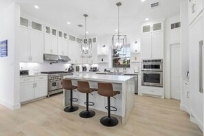 A modern kitchen with white cabinetry, a central island with bar stools, stainless steel appliances, and pendant lighting. - Lux Home-Cinema+GamesGolf-Heated Pool-Hot Tub (Dallas)