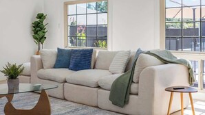 Ein Wohnzimmer mit einer Couch, einem Glastisch, einem kleinen Beistelltisch und einer Topfpflanze.